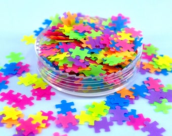 May include: A clear plastic container filled with colorful puzzle-shaped confetti. The confetti is in various shades of pink, blue, green, yellow, and orange. The confetti is scattered around the container on a light blue background.