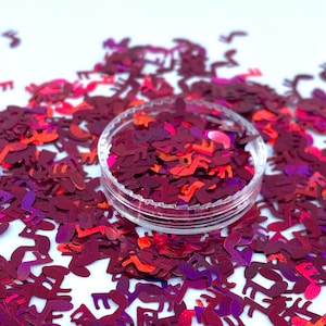 May include: A clear plastic container filled with red and purple iridescent glitter shaped like music notes. The glitter is scattered on a white surface.
