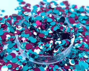 May include: A close-up of a clear container filled with teal and magenta glitter confetti. The confetti is scattered around the container on a white surface.
