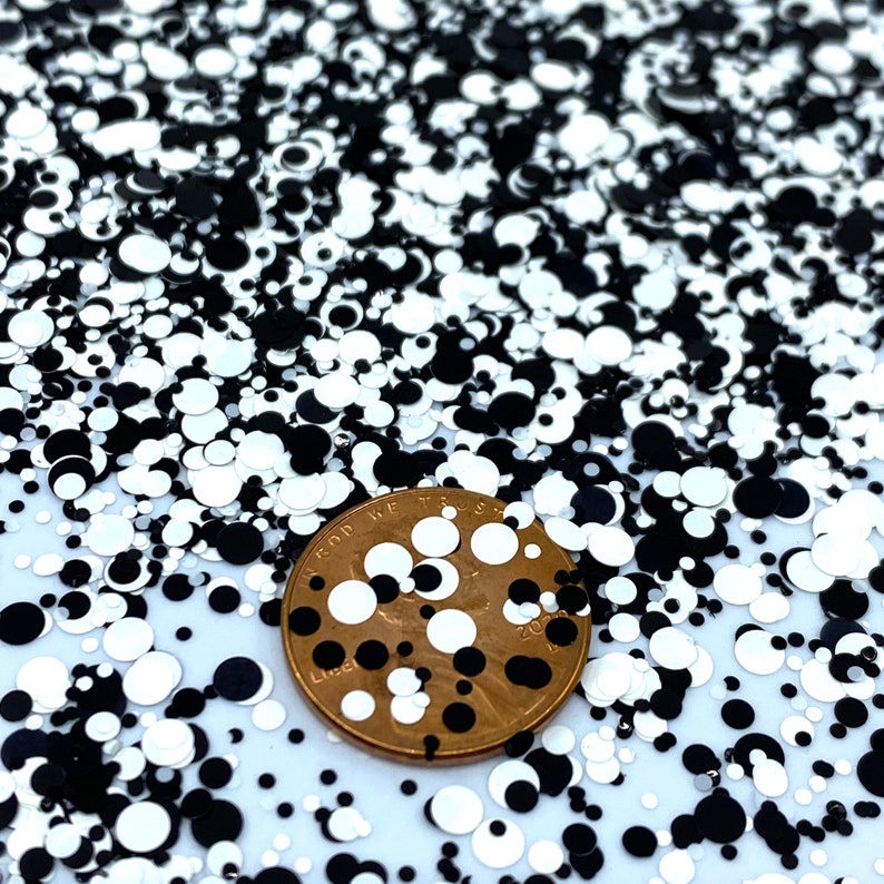 May include: Black and white glitter with a variety of sizes and shapes. A US penny is placed on top of the glitter for scale.