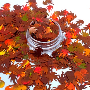 May include: A clear plastic jar filled with flame-shaped sequins in shades of brown, orange, and red. The sequins are scattered around the jar, creating a shimmering effect. The sequins are ideal for nail art, crafts, or other decorative projects.