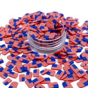 May include: A clear plastic container filled with red, white, and blue American flag-themed sprinkles. The sprinkles are scattered around the container on a white surface.
