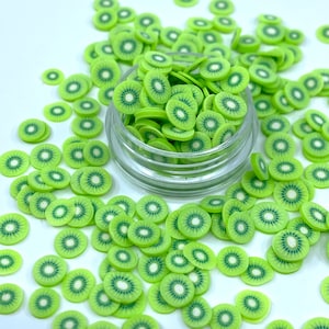 May include: A clear glass jar filled with green and white kiwi fruit slices. The kiwi slices are scattered around the jar on a white surface.