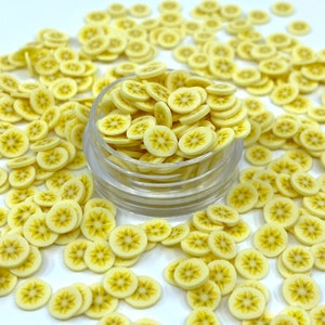 May include: A clear plastic container filled with yellow banana slice shaped sprinkles. The container is surrounded by more sprinkles on a white background.