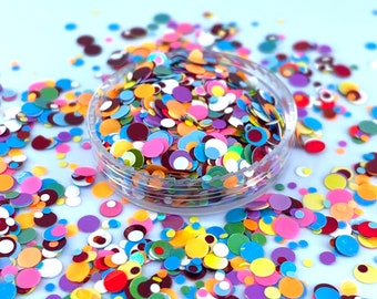 May include: A clear plastic container filled with colorful round confetti. The confetti is a mix of red, orange, yellow, green, blue, purple, and white. The confetti is scattered on a light blue surface.