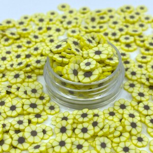 May include: A clear jar filled with small yellow and brown flower-shaped sprinkles. The sprinkles are scattered around the jar on a white surface.