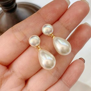 Può includere: Un paio di orecchini pendenti composti da un piccolo perno a perla bianca rotonda collegato tramite un anello in metallo dorato a un ciondolo a perla bianca più grande, liscio e a forma di goccia. Gli orecchini hanno una finitura lucida e simmetrica.
