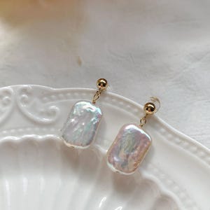 Puede incluir: Un par de pendientes de perlas rectangulares con superficies iridiscentes. Cada pendiente presenta un perno dorado y una delicada cadena dorada que conecta con la perla. Los pendientes se exhiben sobre un plato blanco decorativo.