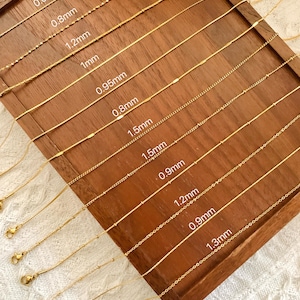 Puede incluir: Un tablero de exhibición de madera con cadenas doradas de diferentes anchos. Las cadenas están etiquetadas con su ancho en milímetros, que van de 0,65 mm a 1,5 mm.