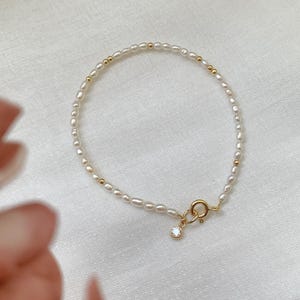 Pode incluir: Delicada pulseira de pérolas com pequenas pérolas ovais e contas douradas. A pulseira tem um fecho dourado e um pequeno pingente de pedra preciosa transparente. A joia é exibida em uma superfície branca texturizada.