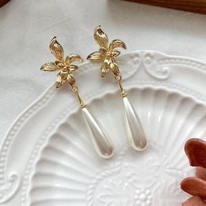 Puede incluir: Un par de pendientes de oro con un diseño floral y una perla blanca en forma de gota. Los pendientes están sobre un plato blanco con un patrón decorativo.