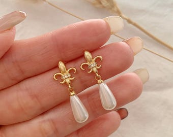 Puede incluir: Un par de pendientes colgantes de tono dorado con un motivo de flor de lis en la parte superior, adornado con pequeños cristales transparentes, y un colgante de perla sintética blanca en forma de lágrima suspendido de la base.