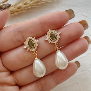 May include: A pair of gold-tone drop earrings featuring an oval-shaped olive green centre stone surrounded by a halo of small clear crystals. Each earring is finished with a smooth, white, teardrop-shaped faux pearl dangling from the base.