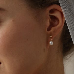 May include: A gold earring featuring a small stud and a hanging white pearl. The earring is worn on an ear. The background is blurred, with a suggestion of a veil.