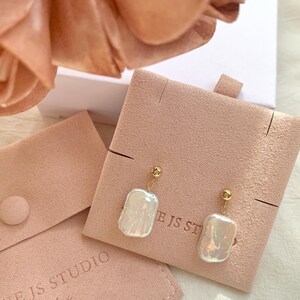 Puede incluir: Un par de pendientes colgantes dorados con grandes perlas barrocas blancas irregulares. Los pendientes se exhiben en una tarjeta de joyer&iacute;a de gamuza rosa. Las palabras "NIE JS STUDIO Jewelry" est&aacute;n impresas en una bolsa rosa.