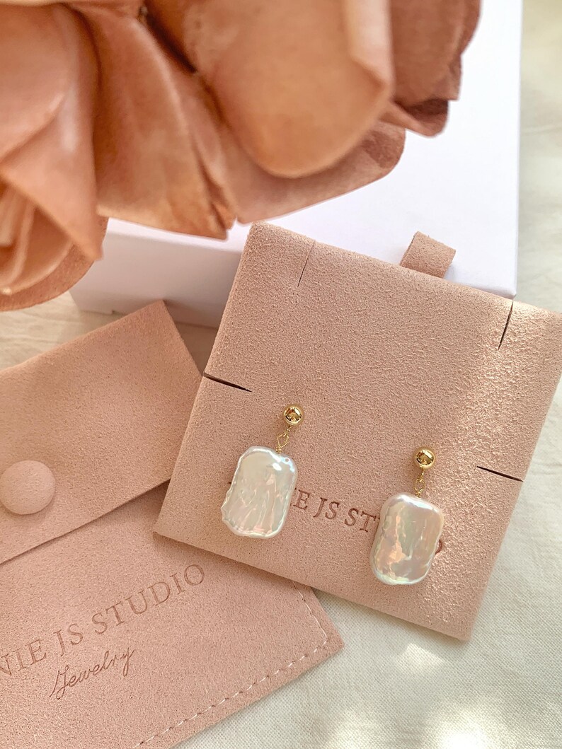 Puede incluir: Un par de pendientes colgantes dorados con grandes perlas barrocas blancas irregulares. Los pendientes se exhiben en una tarjeta de joyer&iacute;a de gamuza rosa. Las palabras "NIE JS STUDIO Jewelry" est&aacute;n impresas en una bolsa rosa.