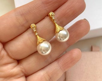 May include: A pair of gold-colored drop earrings with a pearl and diamond design. The earrings have a teardrop shape, with a large, round, white pearl at the bottom and a decorative gold top with small diamonds. The earrings are held in a hand.