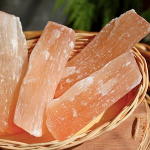 Puede incluir: Cuatro varillas de cristal de selenita naranja en una cesta tejida. Los cristales son translúcidos y tienen una textura estriada.