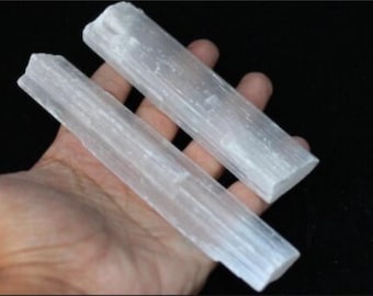 May include: Two white selenite wands, also known as selenite sticks, are held in a hand. The wands are smooth and have a striated texture.