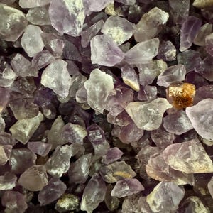 May include: A close-up view of a pile of small, rough, purple amethyst crystals. The crystals are various shades of purple and have a translucent appearance.