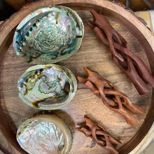 Könnte beinhalten: Drei schillernde Abalone-Muscheln und drei kunstvoll geschnitzte Holzstäbe, die auf einem Holztablett angeordnet sind. Die Muscheln sind hellgrün, blau und violett gefärbt. Die Stäbe sind dunkelbraun.