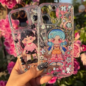 Puede incluir: Dos fundas transparentes para teléfono con personajes de dibujos animados coloridos y detalles de purpurina. La funda de la izquierda presenta un personaje de pelo negro con ropa rosa y la funda de la derecha presenta un personaje de pelo azul con ropa naranja. Ambas fundas tienen la palabra "Love" escrita en ellas.