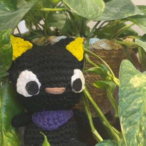 May include: A black crocheted cat plush toy with yellow ears and white eyes. The toy is wearing a purple scarf and is sitting in front of a potted plant.