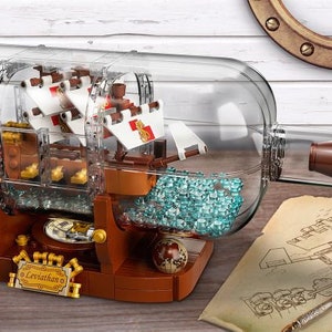 May include: A detailed model ship inside a clear glass bottle. The ship has white sails with red accents and is surrounded by blue-green elements. The bottle sits on a brown wooden base with a gold nameplate that reads "Leviathan".