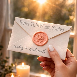 Puede incluir: Un sobre color crema sellado con un sello de cera rosa con un diseño de rosa. El texto en el sobre dice "Read This When You're Wishing For Good Weather". La escena incluye velas encendidas y una ventana con luz solar.