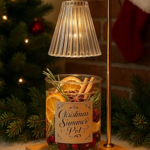 May include: A festive glass jar with the text "Christmas Summer Pot" containing orange slices, cinnamon sticks, and cranberries, lit by a lamp with a ribbed glass shade. The lamp has a gold-coloured metal frame and sits on a wooden base.