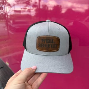 Puede incluir: Gorra de camionero gris y negra con visera curva. La gorra presenta un parche de cuero marrón con el texto "WELL, SHEEYIT" en negrita. La gorra se muestra sobre un fondo rosa.