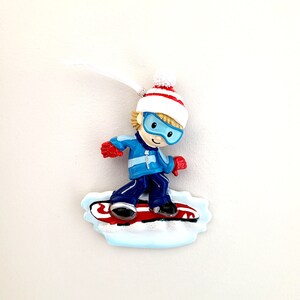 May include: A blue and white ornament of a boy wearing a red and white hat and blue goggles, snowboarding on a red and white snowboard.