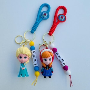May include: Two Frozen character keychains with colorful bead accents. The keychain on the left features Elsa with the name "EMMY" spelled out in white beads. The keychain on the right features Anna with the name "ANNA" spelled out in white beads.