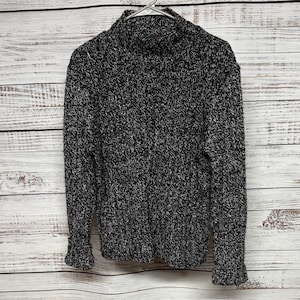 May include: A black and white speckled knit sweater with a mock turtleneck. The sweater is hanging on a metal hanger against a light wood-paneled background. The sleeves have ribbed cuffs.