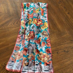 Puede incluir: Bufanda rectangular con estampado floral. La bufanda presenta un fondo turquesa con flores naranjas, amarillas y marrones. Los bordes están ribeteados con un borde color coral. El borde inferior tiene un diseño a rayas blancas y negras.