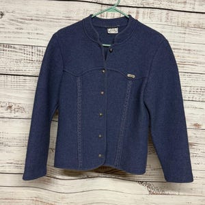 Puede incluir: Chaqueta azul marino con cuello alzado y cierre de botones. La chaqueta presenta costuras decorativas en la parte delantera y un pequeño emblema metálico. Cuelga de una percha verde sobre un fondo de madera.