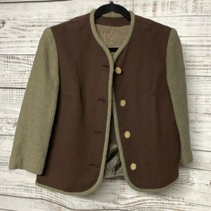 May include: A brown jacket with tan sleeves and trim. The jacket features a round collar and four buttons down the front. The sleeves are a textured tan material, while the body is a smooth brown fabric. The jacket is hanging on a black hanger.