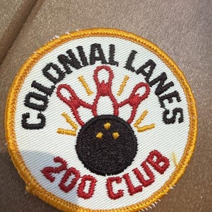 May include: A circular embroidered patch with a white background and a gold border. It features a black bowling ball, red bowling pins, and yellow motion lines. The text reads Colonial Lanes 200 Club in black and red stitched lettering.