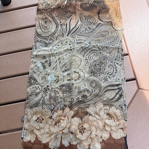May include: A rectangular scarf featuring a brown, tan, and black paisley pattern across the main body. The bottom edge displays a large floral border in cream and brown tones, with the brand name HALSTON printed in white in the bottom corner.
