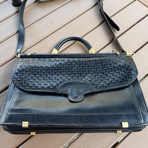 May include: A black leather briefcase featuring a woven texture on the front flap, a top handle, and an adjustable shoulder strap. The bag includes gold-tone metal hardware, a central clasp closure, and four small metal feet on the bottom base.
