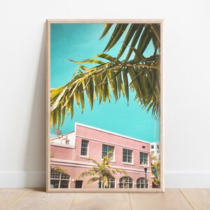 May include: A framed print featuring a pink building with white windows under a bright turquoise sky. Palm fronds frame the top of the image. The print has a light wood frame.