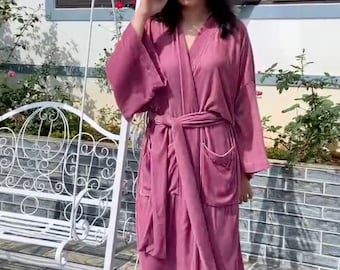 May include: A long, pink velvet robe with a tie belt and pockets. The robe is worn by a person standing on a tiled floor.