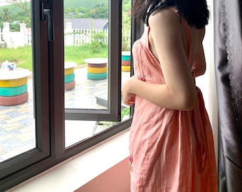 Puede incluir: Una mujer con un vestido rosa se para junto a una ventana mirando hacia una vista de un área verde con árboles y una valla blanca. La ventana está abierta y hay tres mesas de neumáticos coloridos afuera.