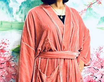 May include: A dusty rose velvet robe featuring long sleeves, a shawl collar, and a matching tie belt at the waist. The garment includes two front patch pockets and is constructed from a soft, plush fabric with a subtle sheen that catches the light.