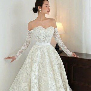 Puede incluir: Vestido de novia blanco, con hombros descubiertos, mangas largas de encaje y falda amplia. El vestido presenta un escote coraz&oacute;n y un cintur&oacute;n blanco. La novia lleva pendientes de perlas y el pelo recogido en un mo&ntilde;o.