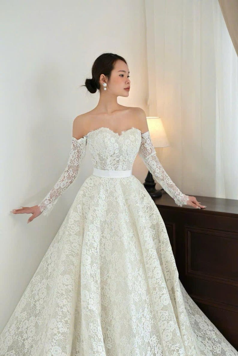 Puede incluir: Vestido de novia blanco, con hombros descubiertos, mangas largas de encaje y falda amplia. El vestido presenta un escote coraz&oacute;n y un cintur&oacute;n blanco. La novia lleva pendientes de perlas y el pelo recogido en un mo&ntilde;o.
