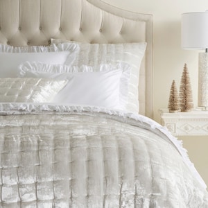 May include: A cream-colored velvet quilt with a square-stitched pattern and ruffled edges, paired with matching square shams and plain white rectangular pillows. The bedding set is displayed on a bed with a tufted, upholstered headboard.