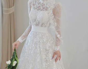 May include: A white wedding dress with long sleeves and a high neckline, adorned with intricate lace and sequin details. The dress features a fitted bodice, a wide white belt, and a flowing skirt. A matching veil and a bouquet of white flowers complete the bridal look.