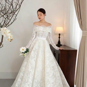 Puede incluir: Vestido de novia blanco, con hombros descubiertos, mangas largas y falda amplia. El vestido est&aacute; hecho de encaje delicado y presenta un cintur&oacute;n de cinta blanca. Un ramo de flores blancas se sostiene en la mano.