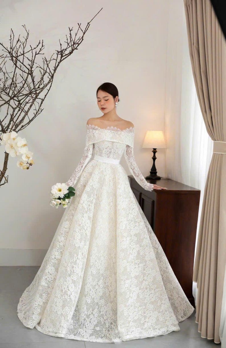 Puede incluir: Vestido de novia blanco, con hombros descubiertos, mangas largas y falda amplia. El vestido est&aacute; hecho de encaje delicado y presenta un cintur&oacute;n de cinta blanca. Un ramo de flores blancas se sostiene en la mano.